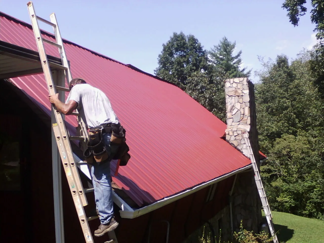 Roofer on a ladder beside a red metal roof and stone chimney — Soffit & Fascia Repair by Roofing Brothers in Saint David